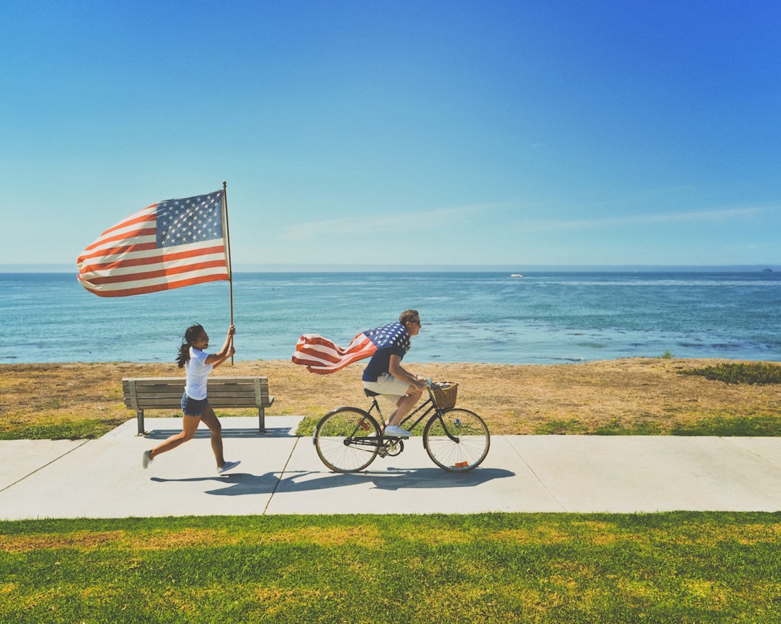 A man rides on a bike with an American flag draped over his shoulders while a woman runs after him holding an american flag