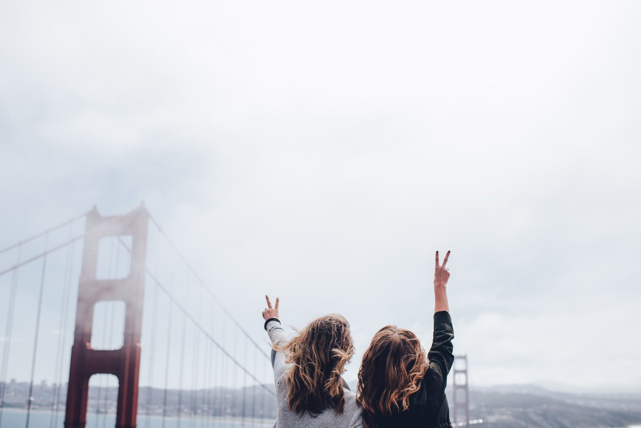 Two women stand in front of the San Francisco bridge, holding up peace signs