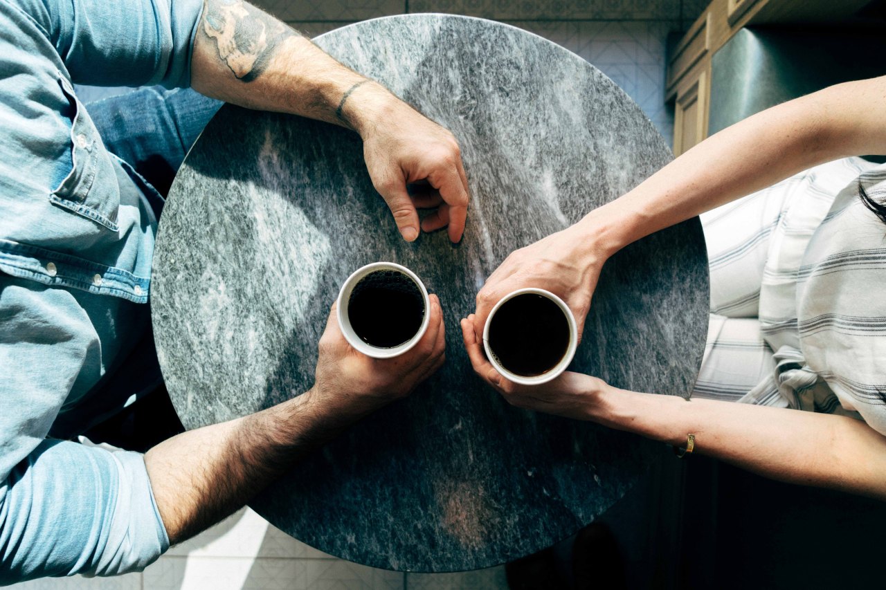 A man and a woman sit across from each other at a table, holding coffee