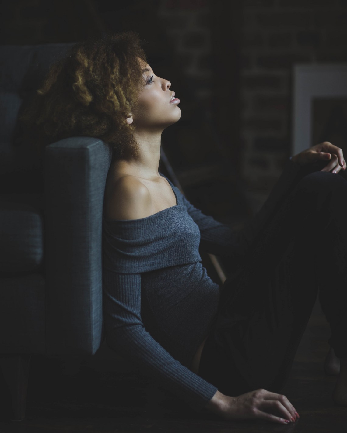 Pretty young curly haired woman sirring on the floor leaning on a gray sofa