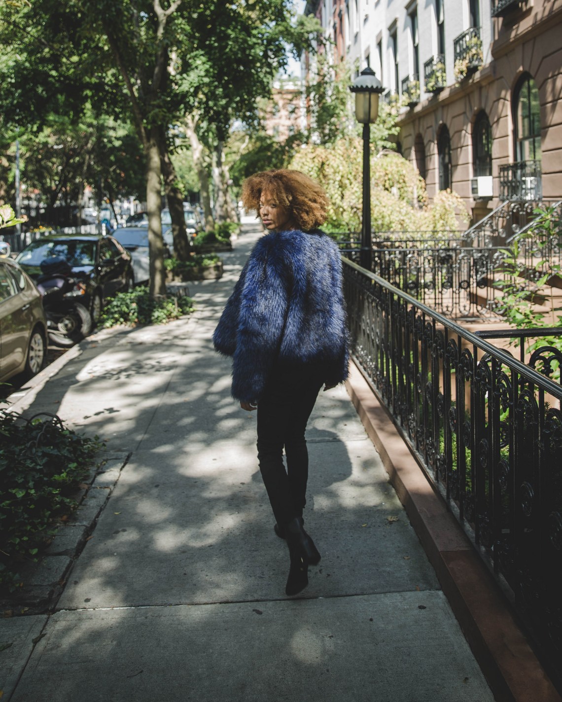 Woman with curly hair in a blue fur jacket looking back as she walks down the pavement