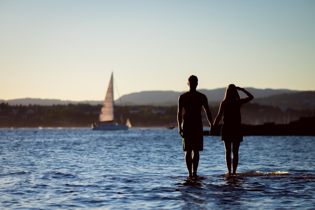 Man and woman standing in the bay watching a sailboat pass by