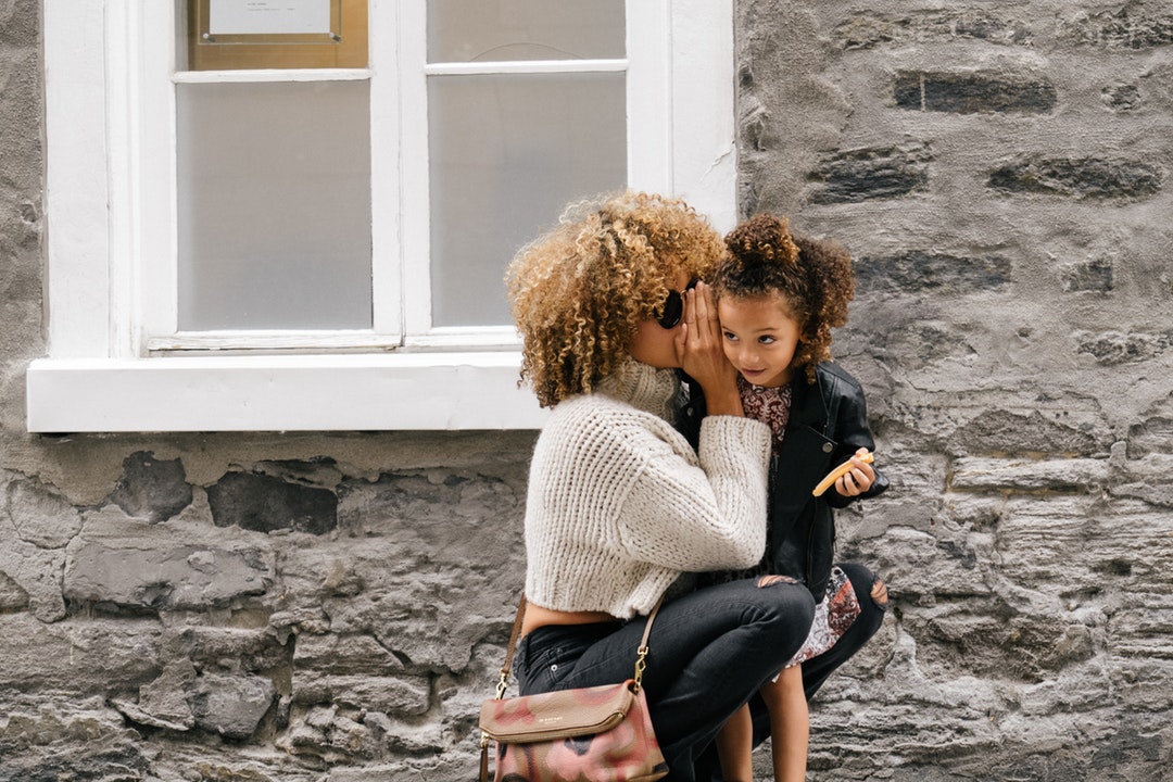 Mother with frizzy hair crouches down to tell daughter a secret on old city street