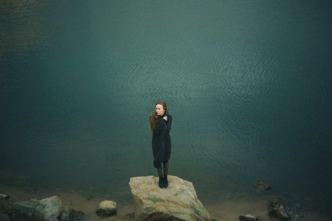 A redheaded woman bundled in a black sweater and black boots, standing on a rock next to a lake