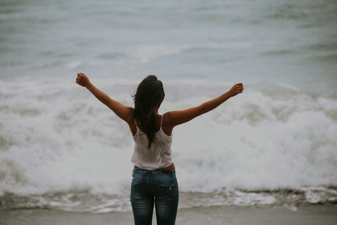 A woman wearing jeans and a white shirt staring out at the ocean with her arms up at Bodega Bay