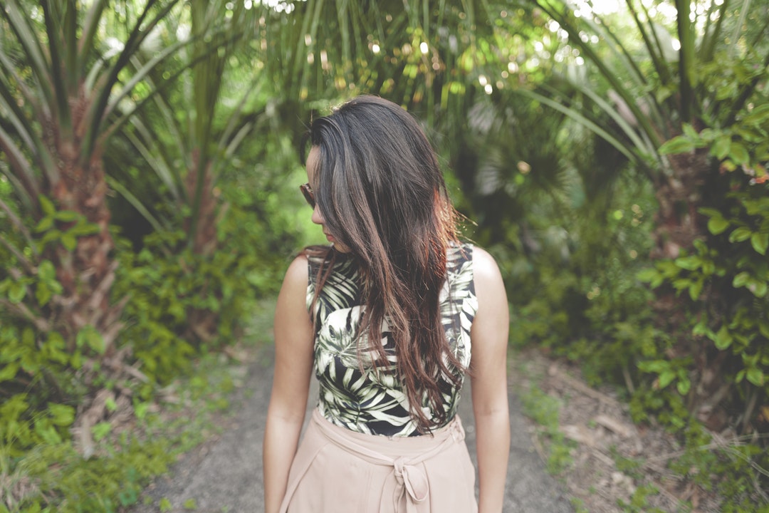 A young dark-haired woman in sunglasses tilting her head to the side in a tropical setting