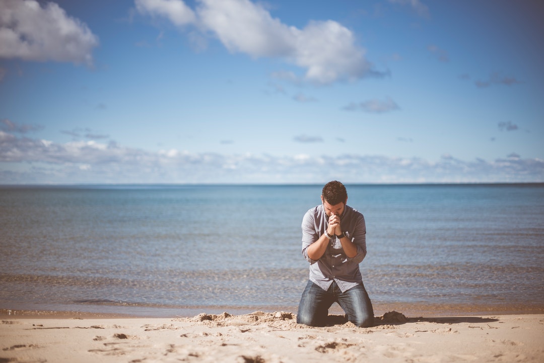 A man in a shirt and jeans kneeling on the edge of a beach with his hands clasped against his chin