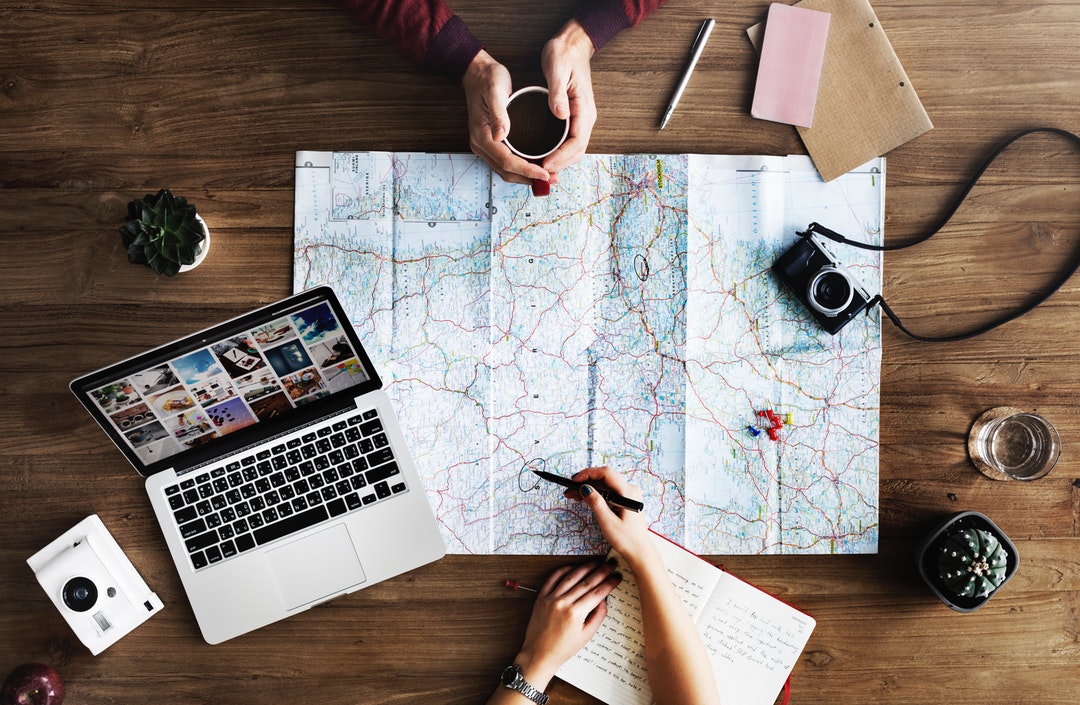 An overhead shot of two people planning a trip with a map and a laptop on a wooden surface