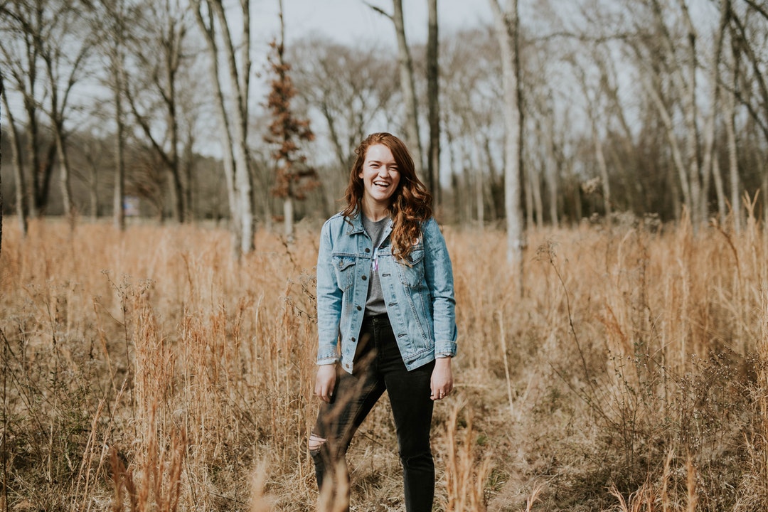 A woman in a denim jacket laughs in a field of long brown grasses and trees