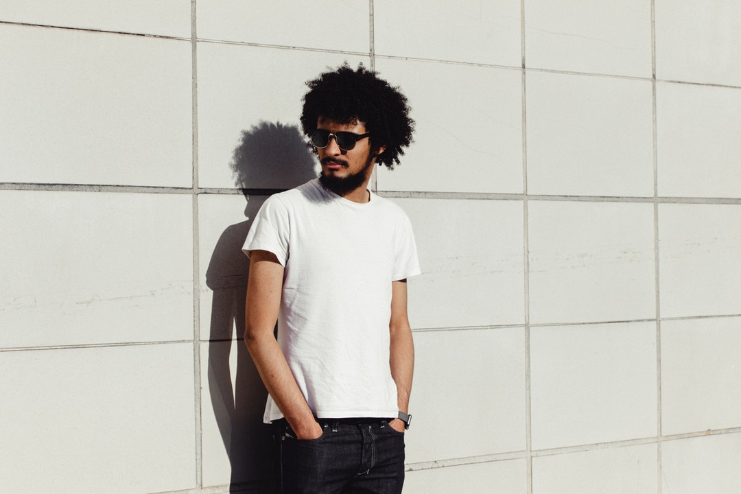 Man with curly hair wearing sunglasses standing against a wall