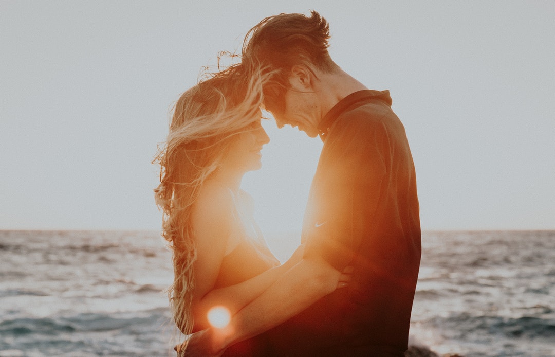 Young man and woman stand to frame the setting sun behind them over the sea