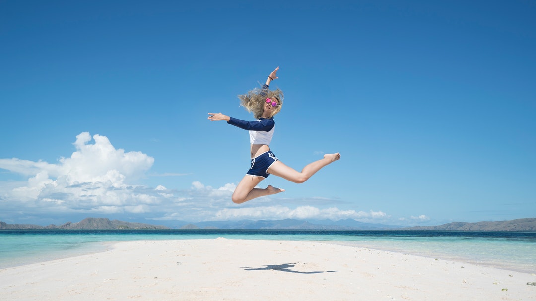 A young woman jumping high above a patch of white sand surrounded by the turquoise sea