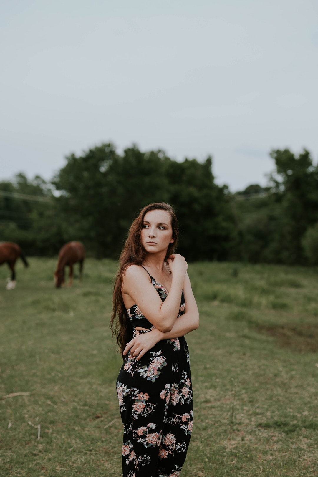 A young woman in a dress standing on a green pasture with horses grazing at the back