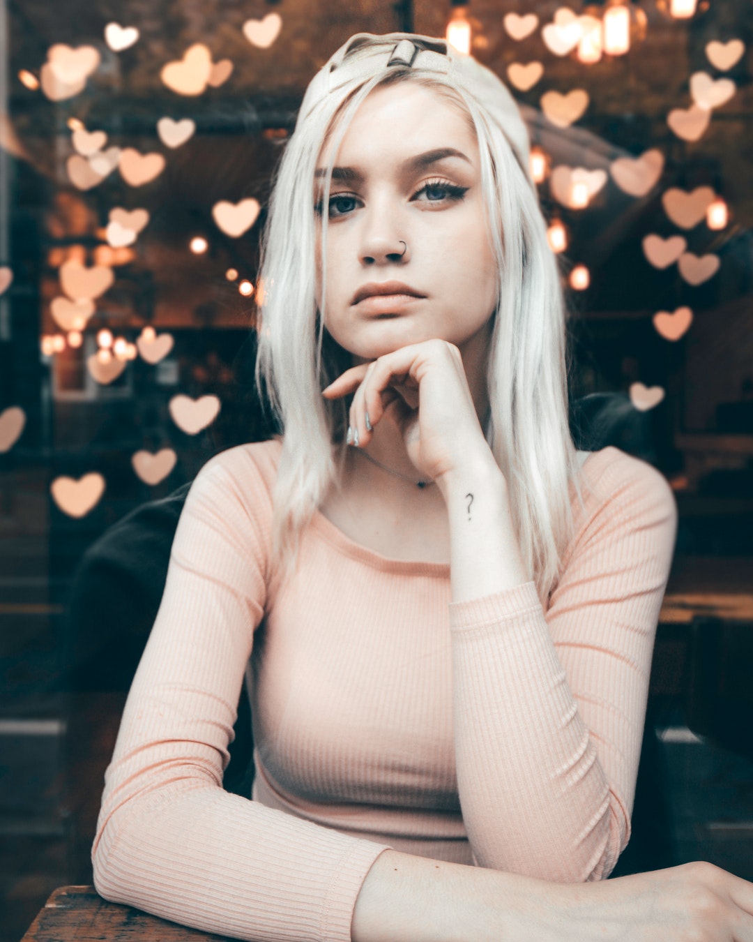 A blonde woman sitting at a table looking at the camera with paper hearts hanging from the ceiling behind her