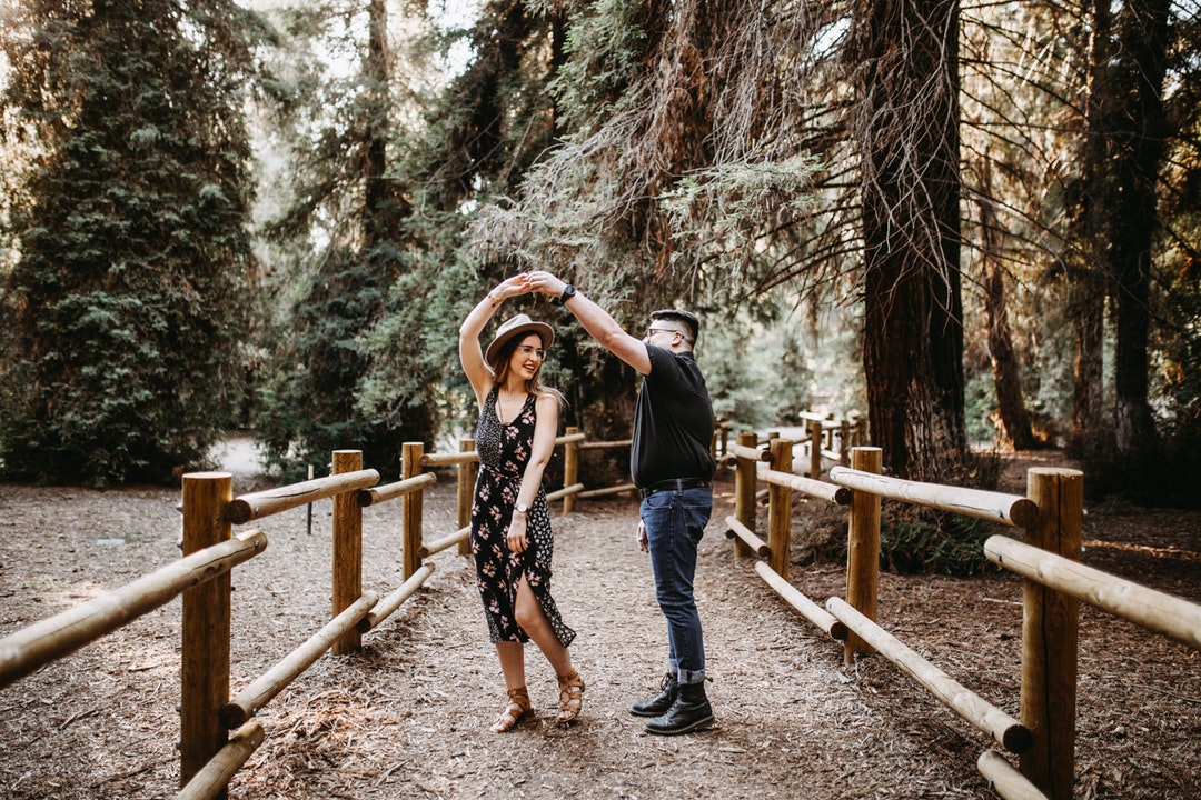 A man and a woman dance, holding hands, on a fenced-in pathway in the woods