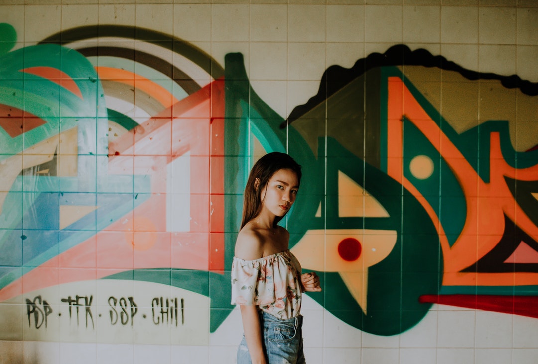 A young woman in an off-shoulder top looks at the camera against a graffiti wall.