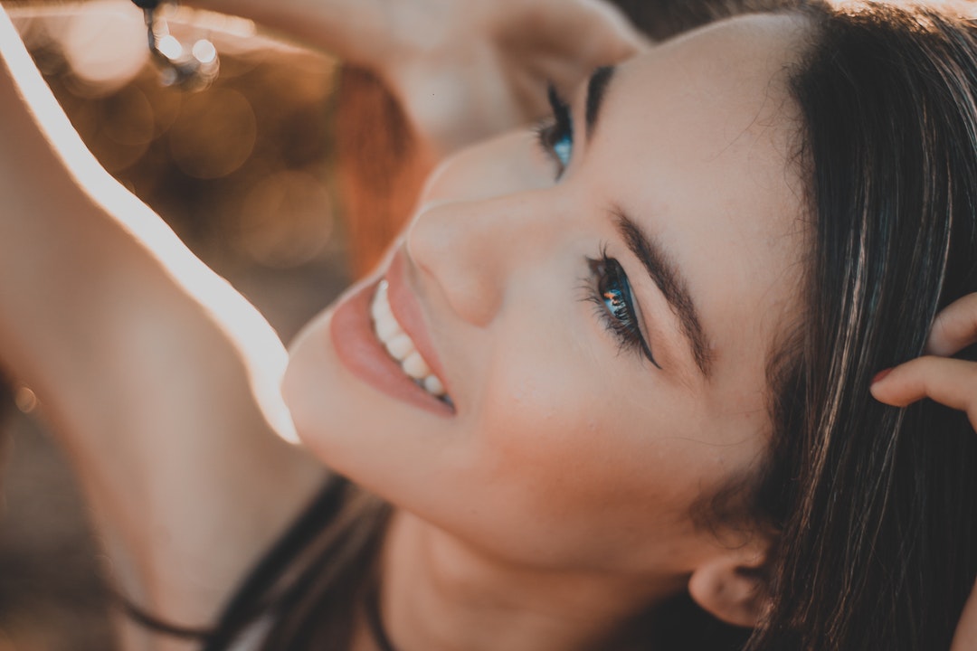 A woman in black eyeliner looks upward and smiles with her hands in her hair