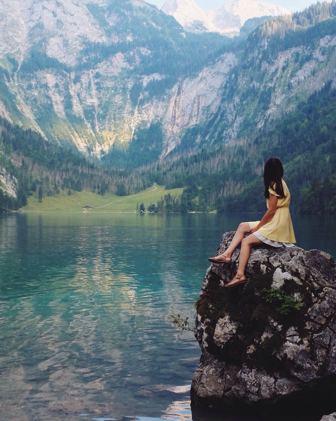 A woman in a yellow dress sitting on a large rock next to a lake and mountains in Шёнау-ам-Кёнигсзе, Бавария, Германия