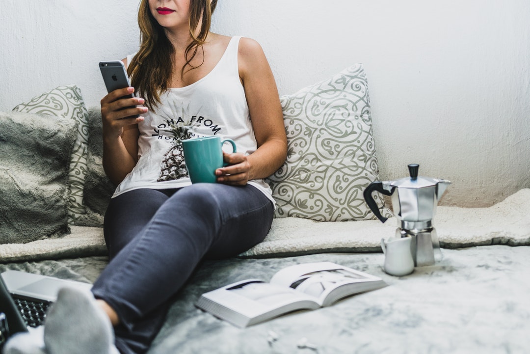 A woman sits on a bed with an open book, holding a mug and her cell phone