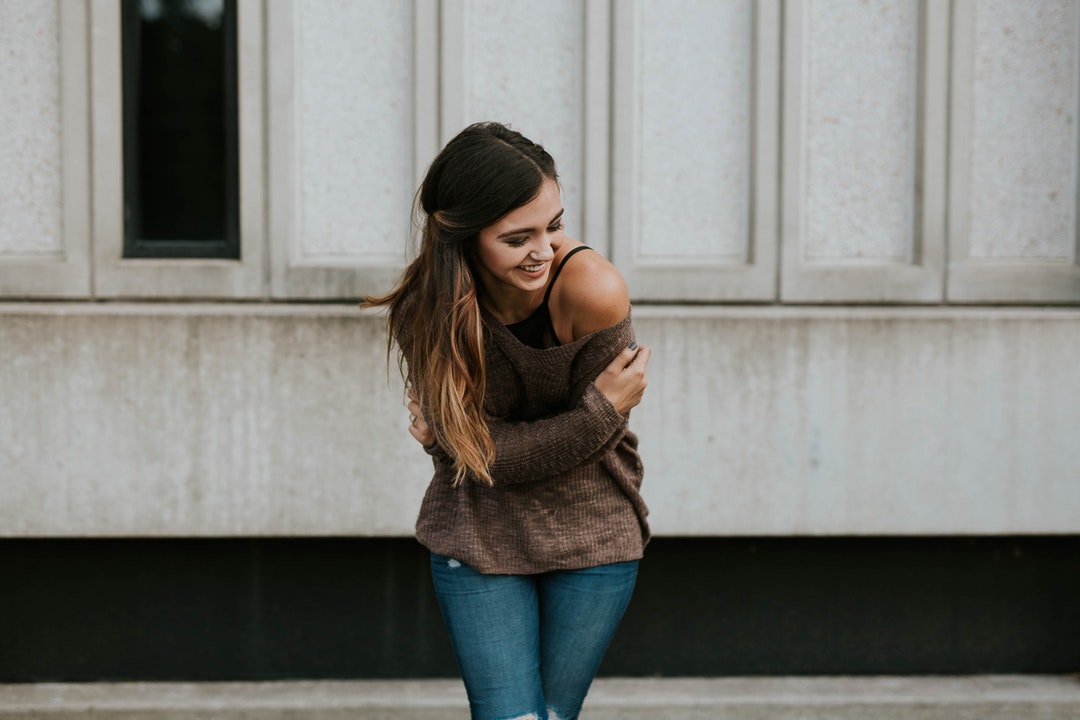 A woman in an off-the-shoulder sweater leans forward while laughing in front of a stone wall