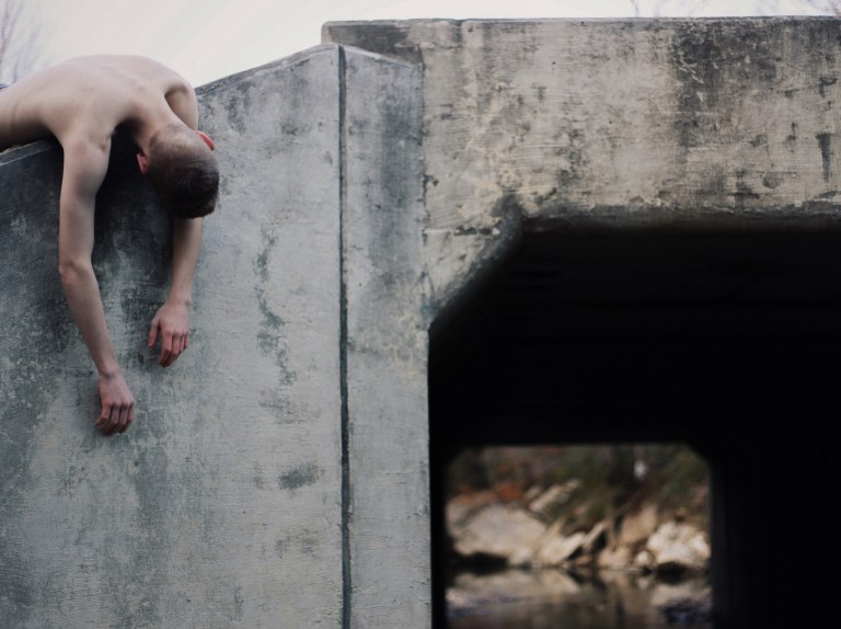 Shirtless man draped over concrete wall of bridge, possibly creepy