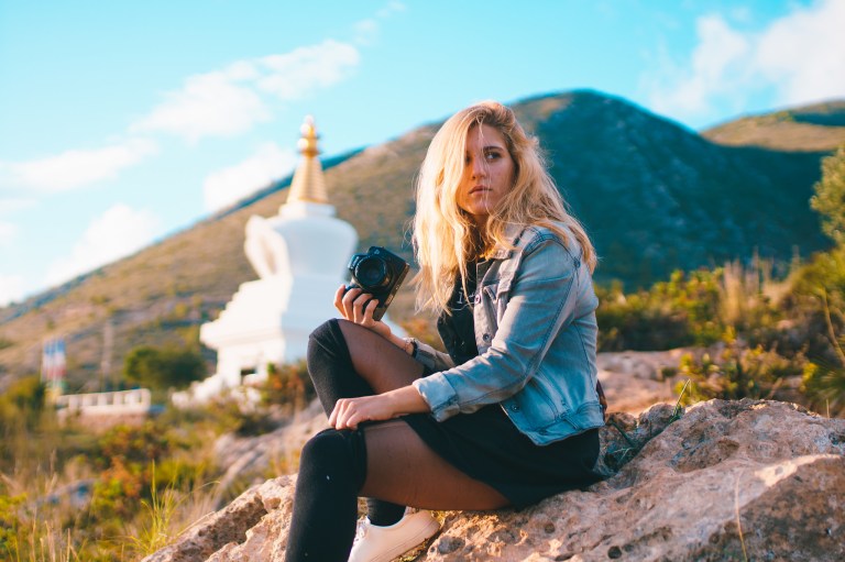 Blonde woman with camera contemplating her next photo op, sitting on a rock