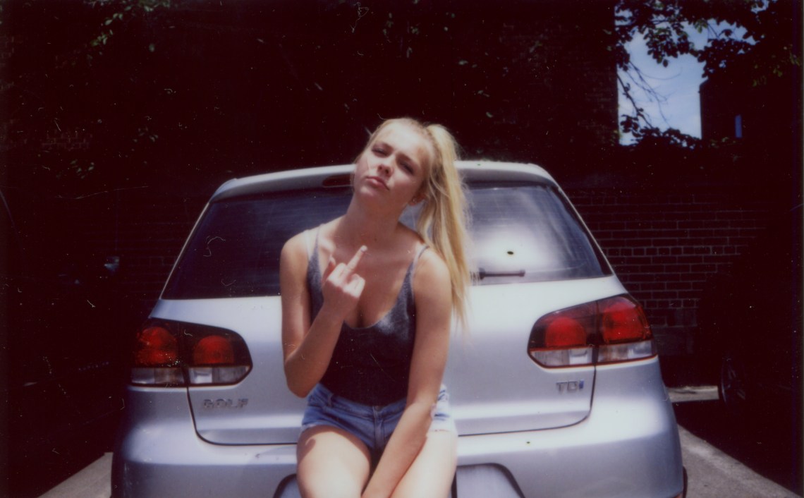 young blonde woman with an attitude flipping someone off, middle finger, leaning on car