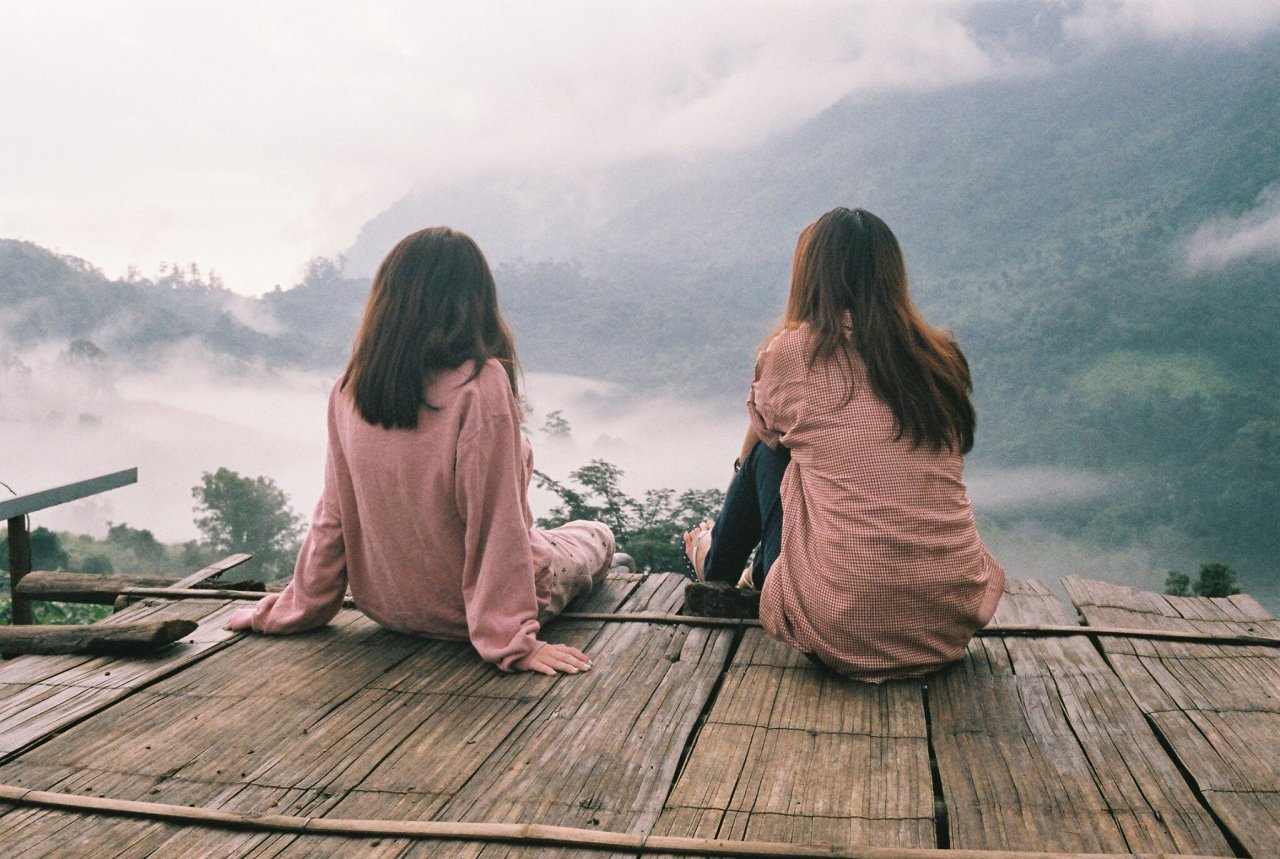 Two brunette girls in matching dusty rose pink shirts sitting on a rickety wooden platform overlooking misty mountains