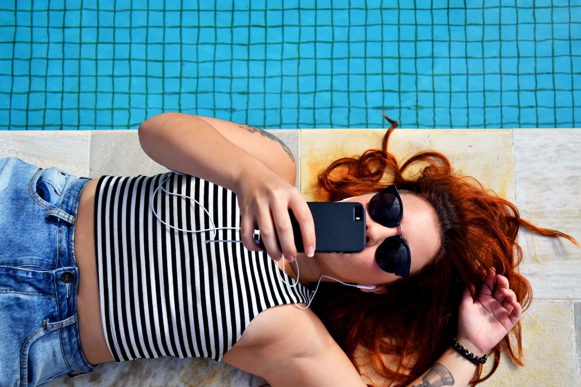 A woman lies by the pool, earphones in her hears, and texts on her iPhone