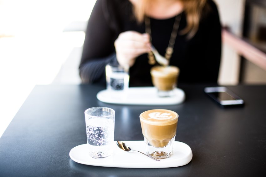 A woman sits at a table with coffee and water, looking down at her phone