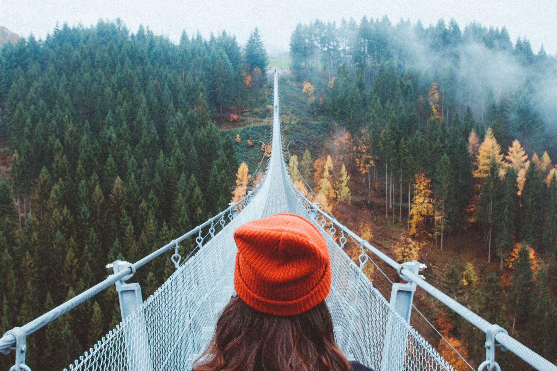 woman wearing knit cap walking on white bridge between trees during daytime