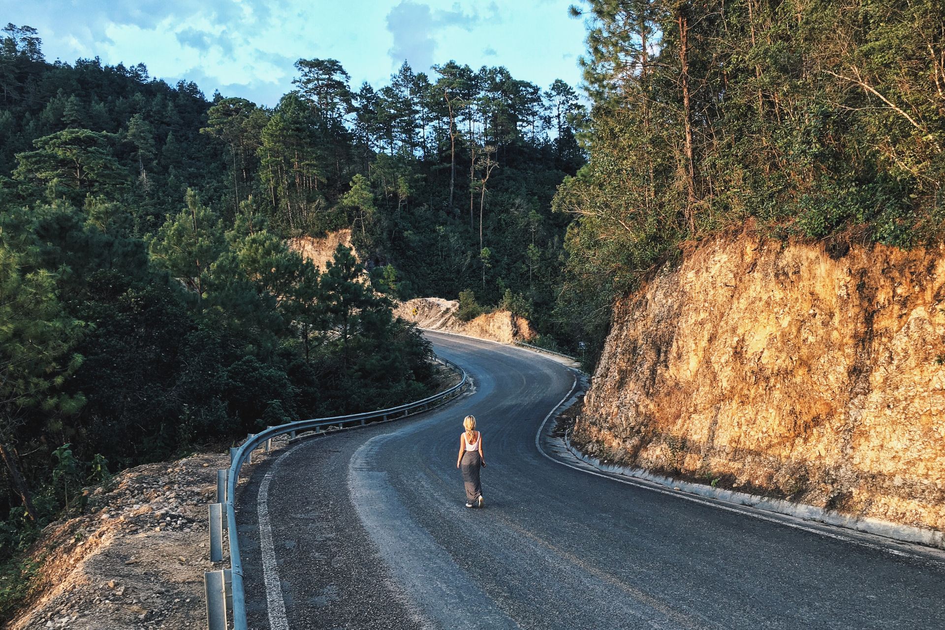 woman walking on gray asphalt road in between trees and rock formations