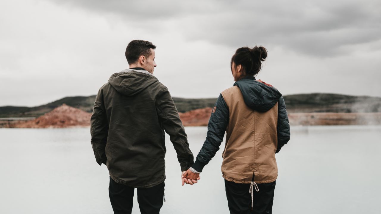 couple holding hand front of body of calm water with mountain distance