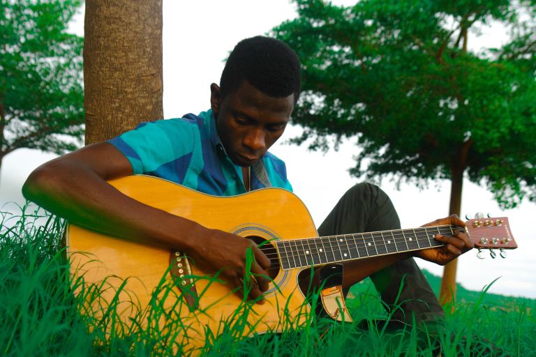 man playing natural finish single-cutaway acoustic guitar while sitting on green grass near brown tree trunk during daytime