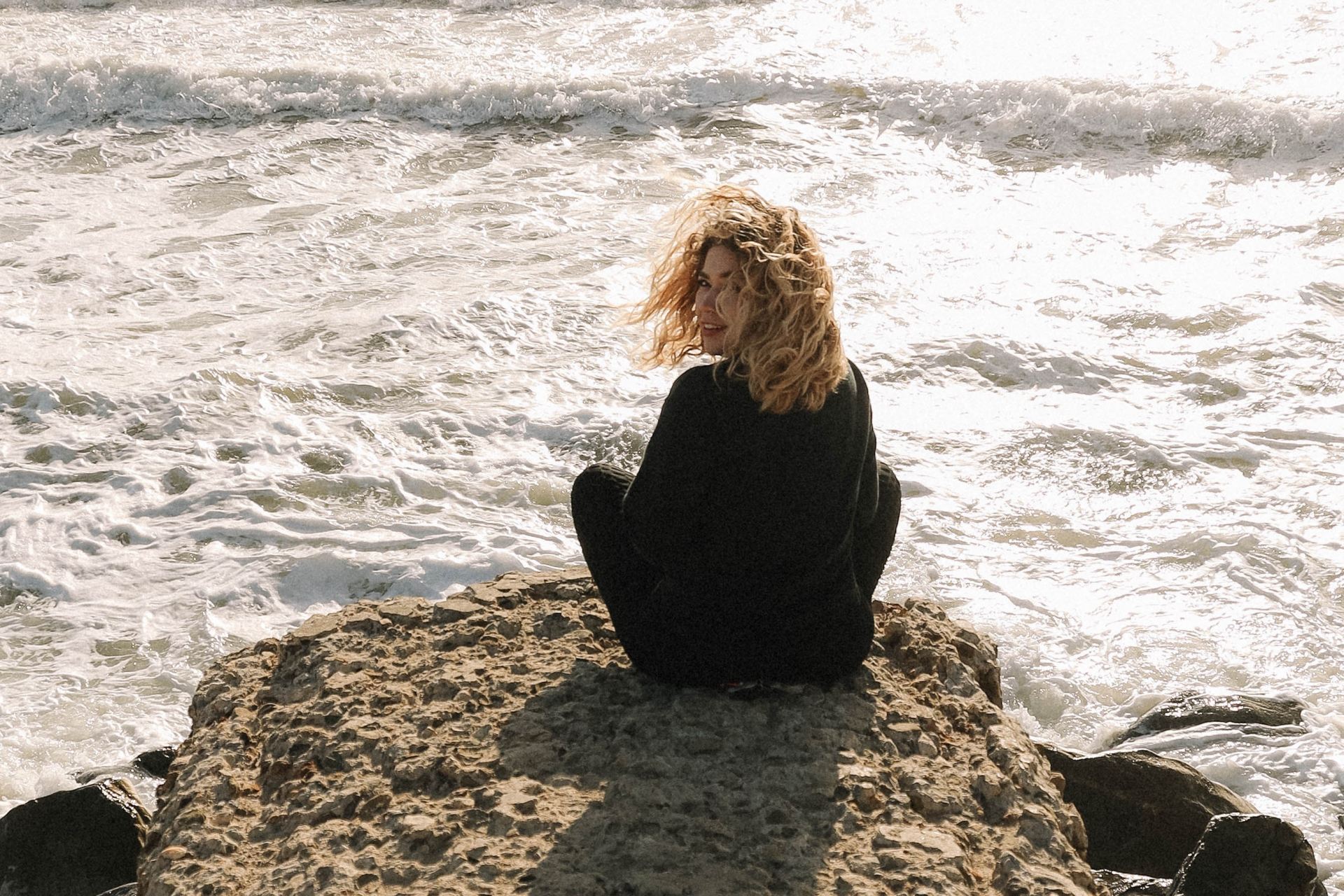 woman in black long sleeve shirt sitting on brown rock near sea during daytime