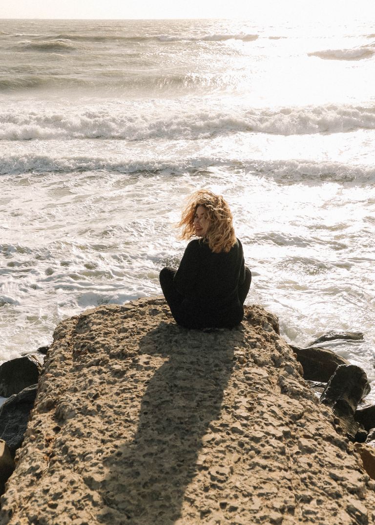 woman in black long sleeve shirt sitting on brown rock near sea during daytime