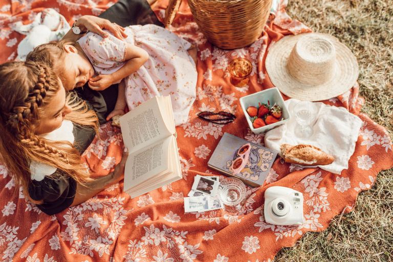 woman reading with girl while lying on orange and white floral picnic mat