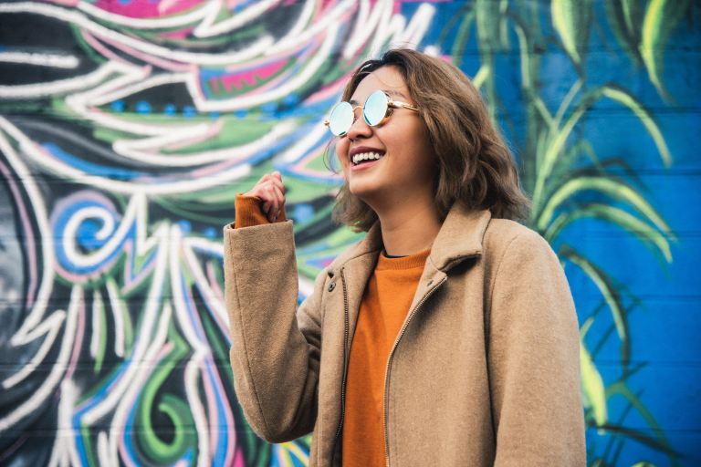 woman wearing orange shirt and beige coat wearing sunglasses while standing near graffiti wall smiling