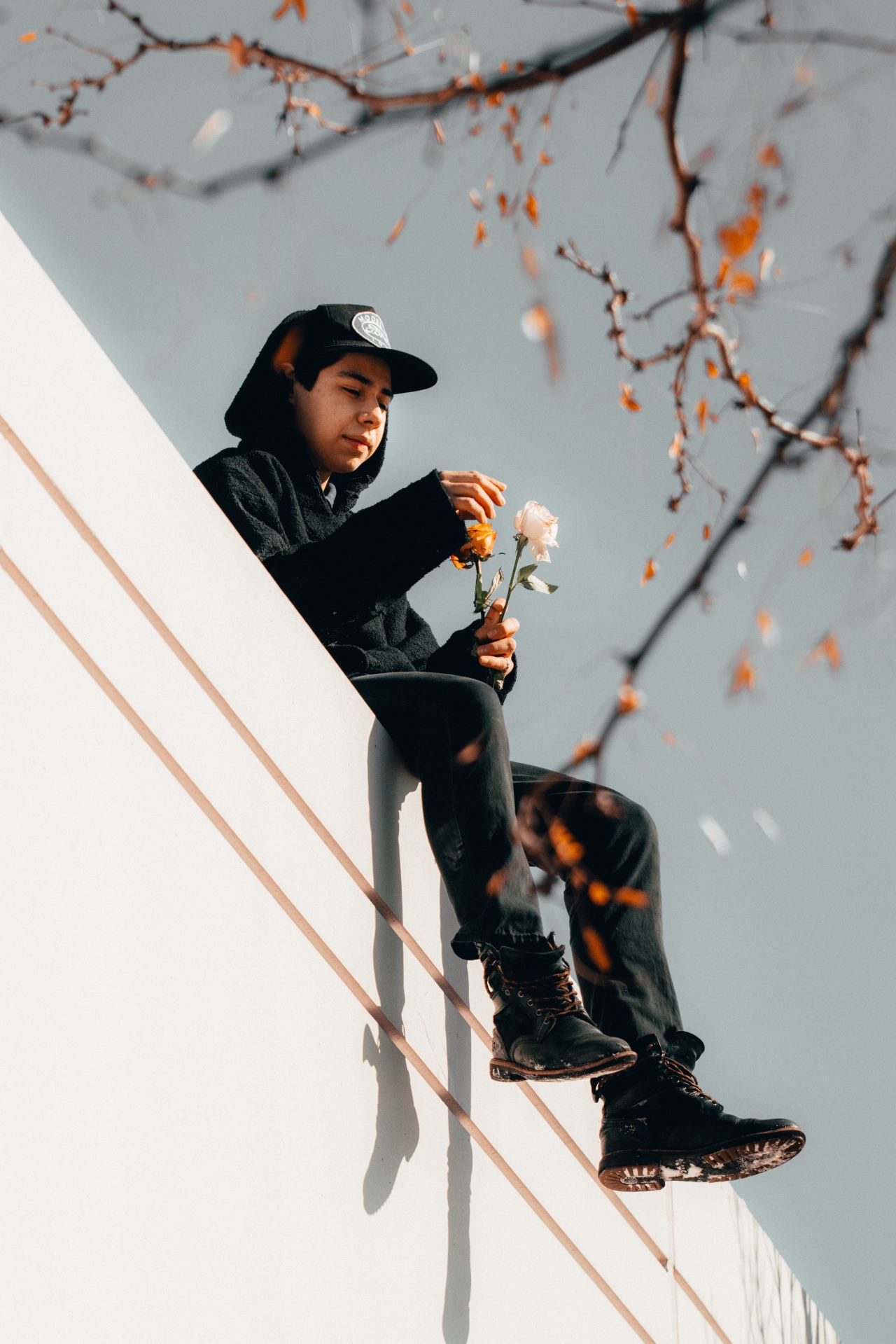 man in black and orange jacket and black hat sitting on white concrete wall during daytime