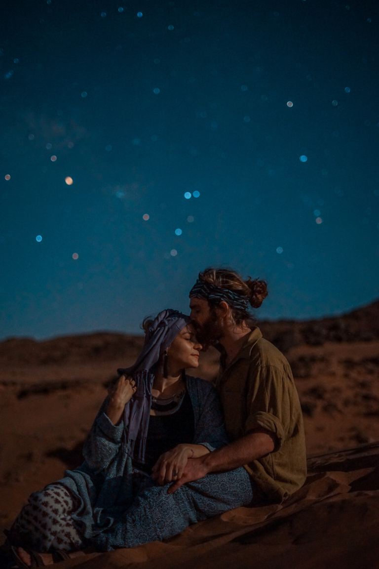 man and woman sitting on desert sand under blue sky during nighttime