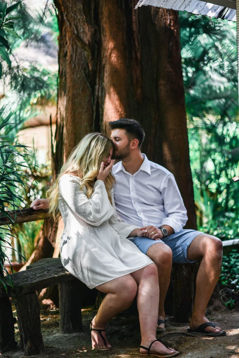 man in white dress shirt kissing a woman in white long-sleeved dress while sitting on bench beside brown large tree at daytime