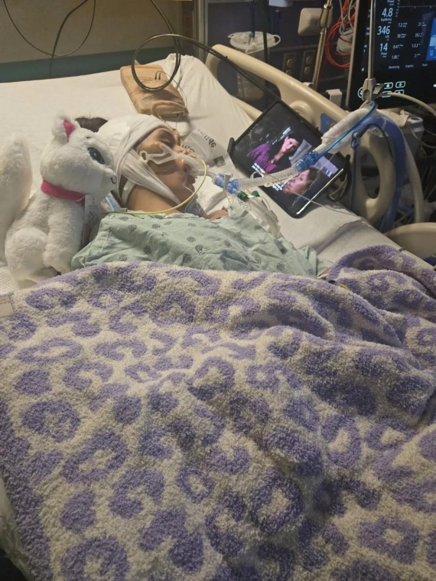 A young girl lies in a hospital bed with medical tubes, a head wrap, a white stuffed bunny beside her, and a tablet mounted nearby.