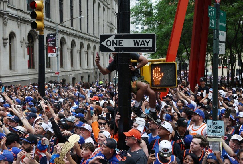 Dense crowd of Knicks fans in blue and orange gear filling a Manhattan street, with one person climbing a traffic sign pole during the Knicks parade.