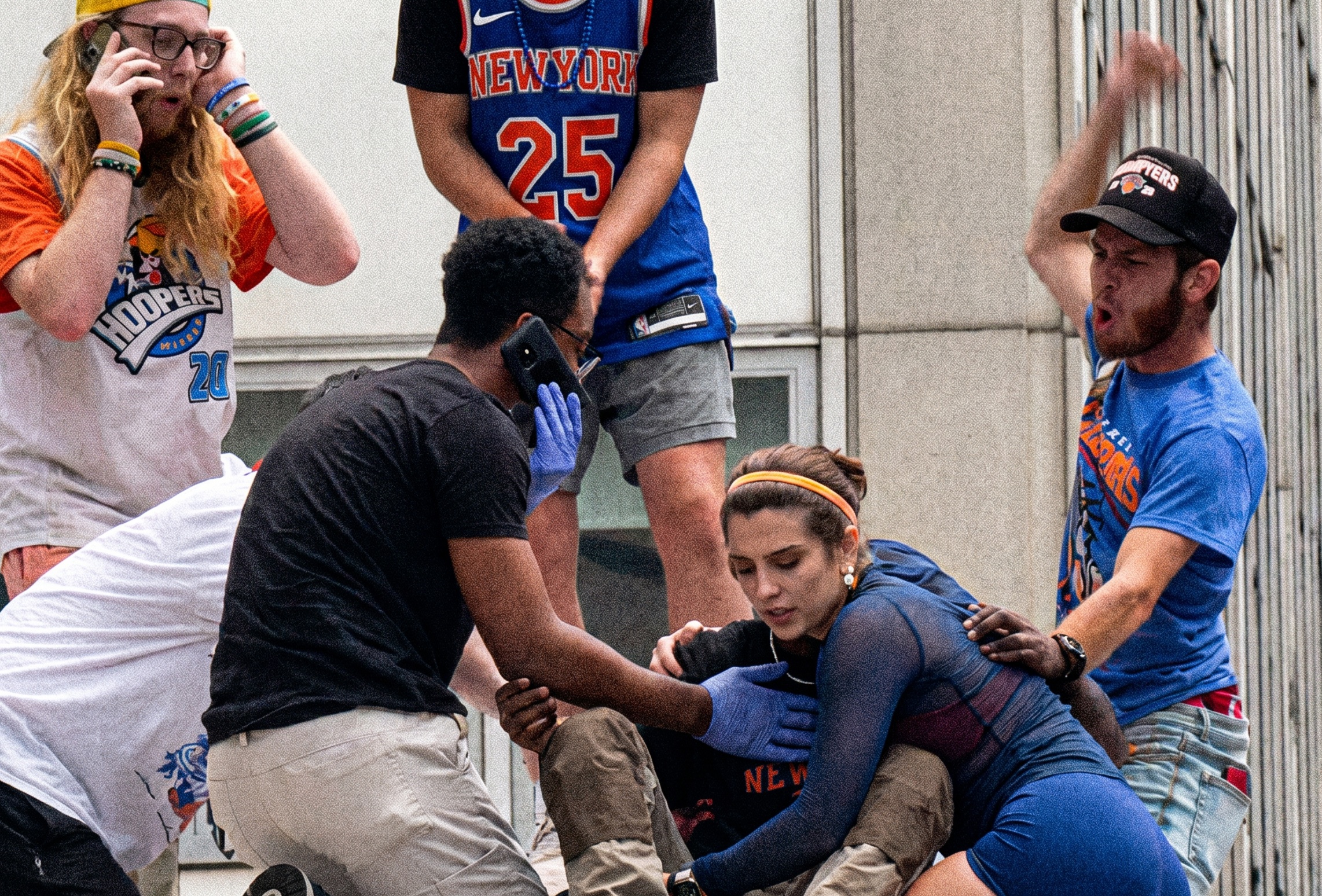 Several people crouch to help a woman in a blue top on the ground, surrounded by bystanders in New York Knicks jerseys.