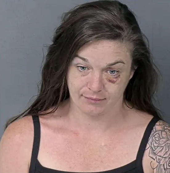 Click to open the image gallery Booking mugshot of a woman with dark hair, blue eyes, and a bruise under her left eye, a tattoo visible on her shoulder.