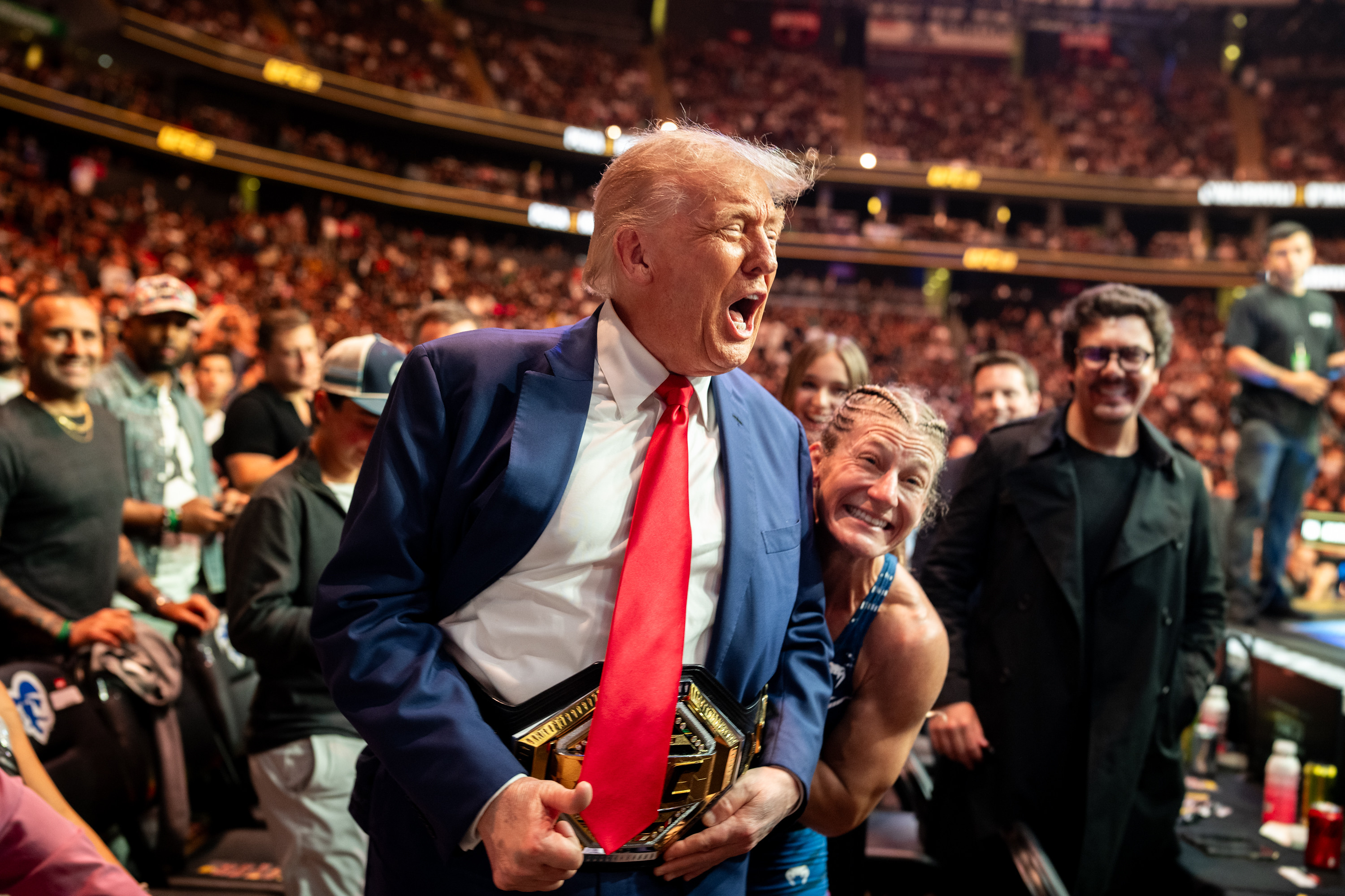 Man in blue suit and red tie holding a UFC championship belt, mouth open in excitement, with a smiling woman beside him in a packed arena.