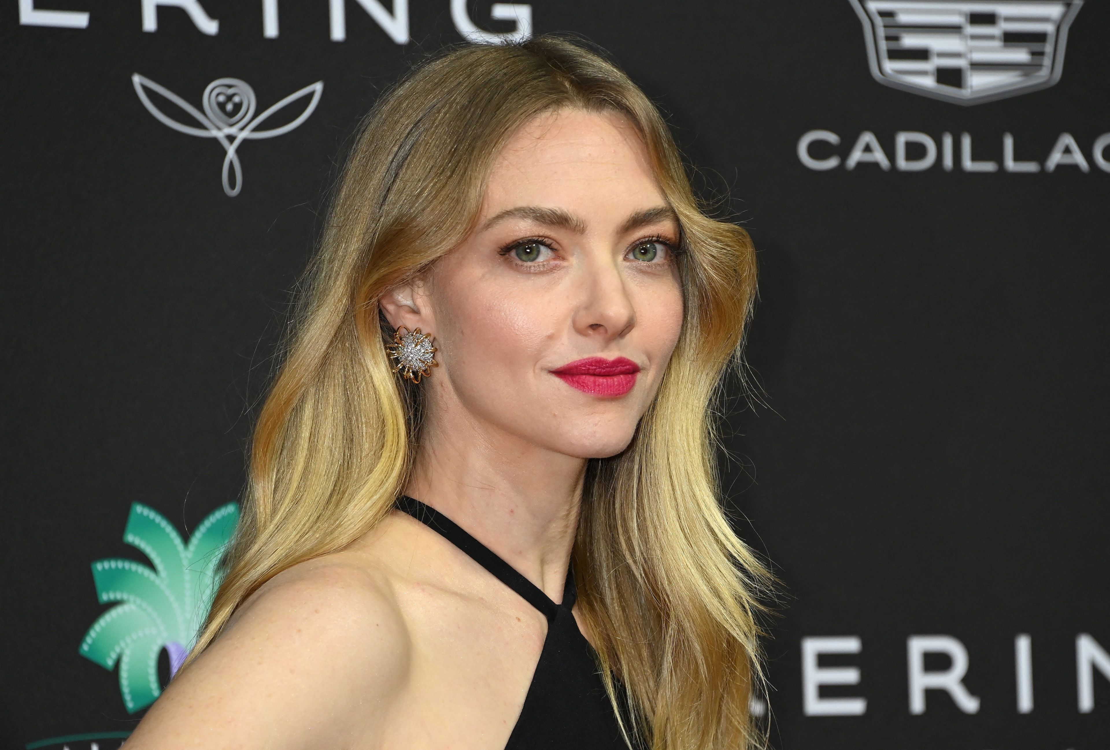 Blonde woman in a black halter dress with red lipstick and a jeweled earring poses at a black-and-teal step-and-repeat backdrop with Cadillac branding.