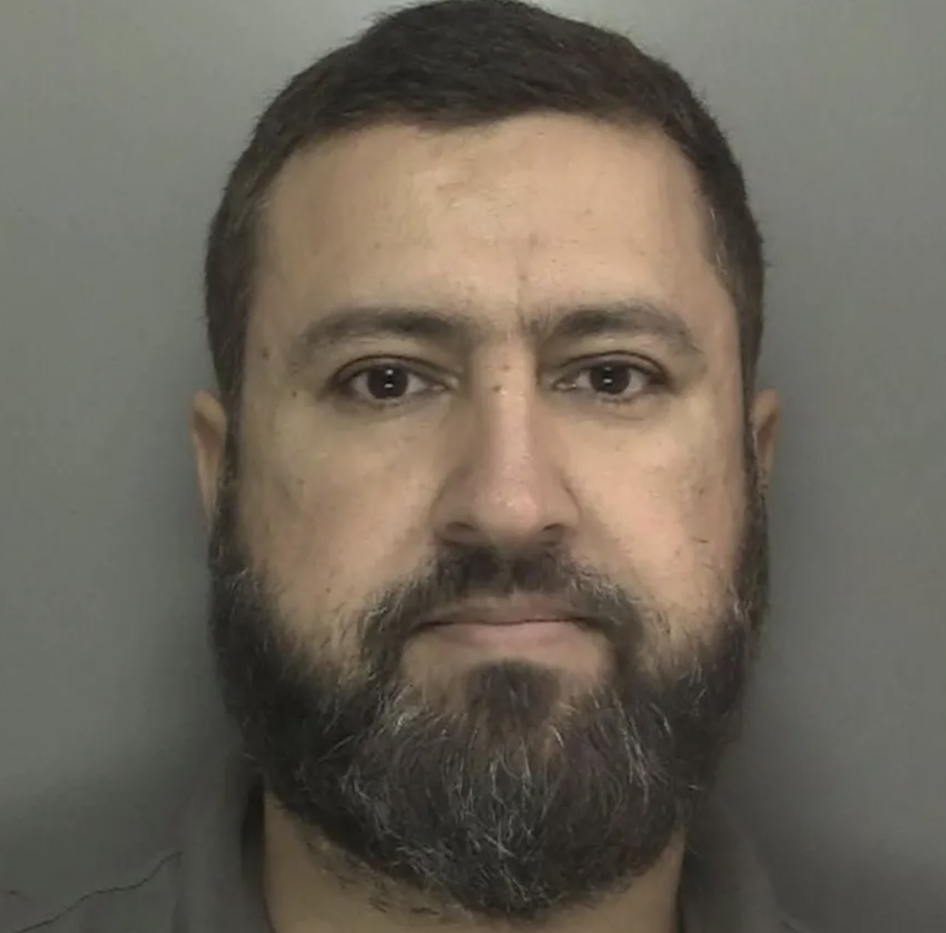 Police mugshot of a middle-aged man with dark hair and a full beard against a grey background.