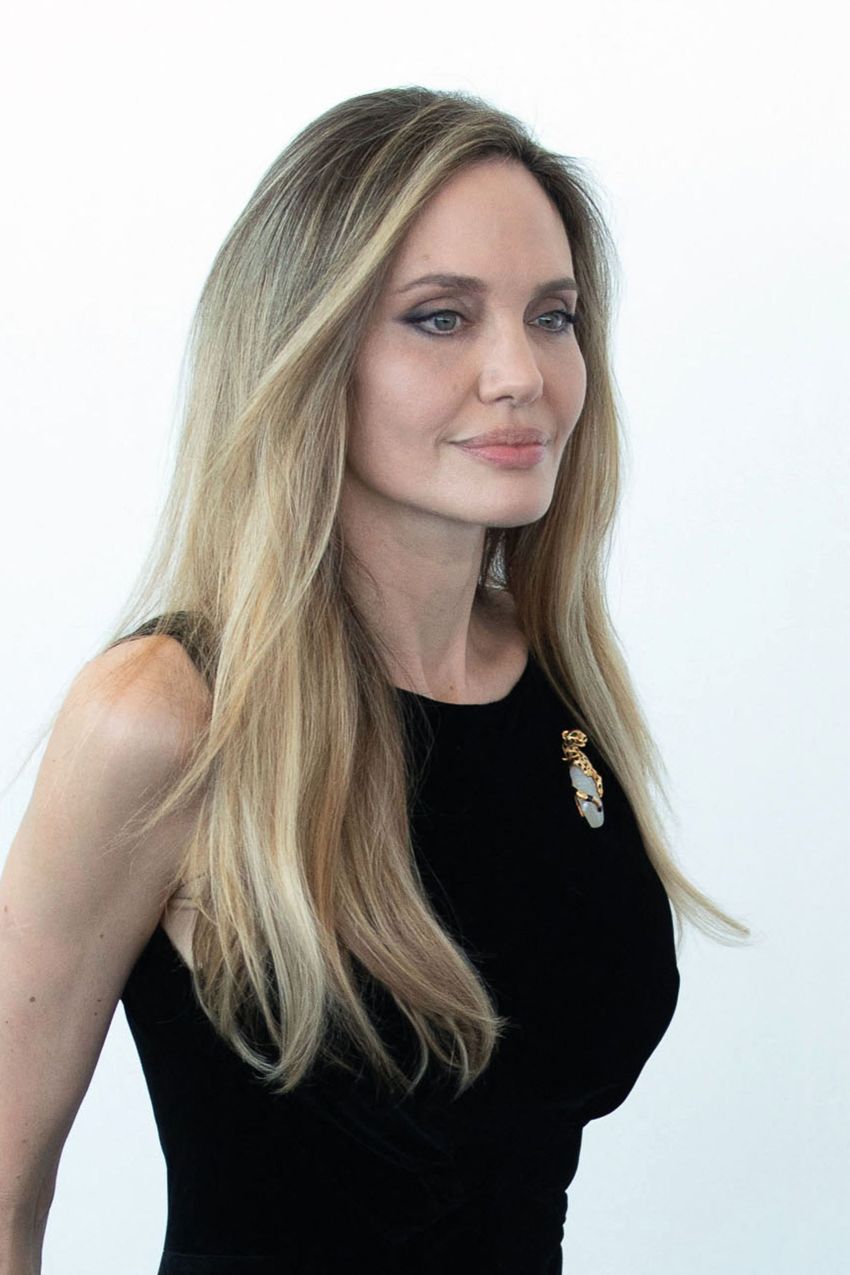 Angelina Jolie in a black sleeveless dress with a gold brooch, long blonde hair, gazing to the side against a white background.
