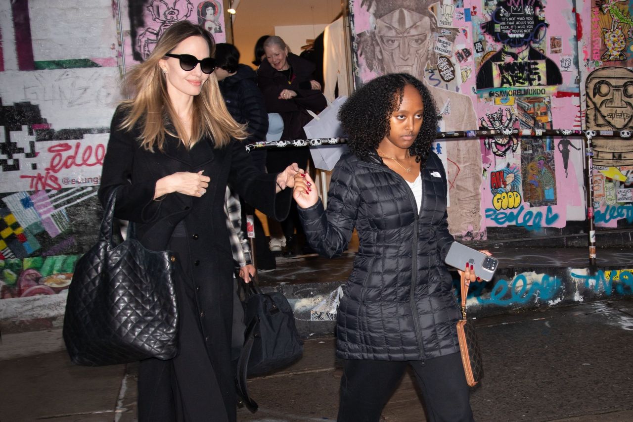 Woman in black coat and sunglasses holding hands with a teenage girl in a North Face puffer jacket walking past a graffiti-covered wall in NYC.